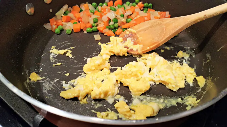 eggs being scramble in half a pan and carrots, onions, and peas in the other half with wooden spoon