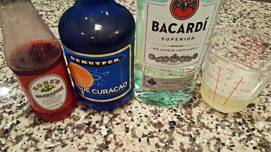 bottle of grenadine, bottle of blue curacao, bottle of bacardi, measuring cup with lemonade in it