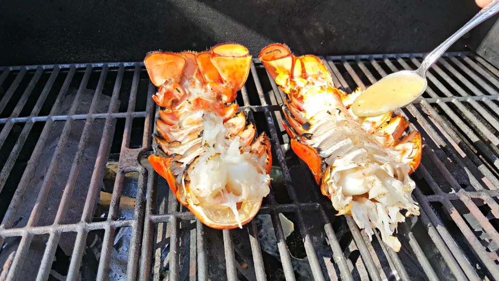 two lobster tails upside down on a gril and a spoon drizzling on some butter