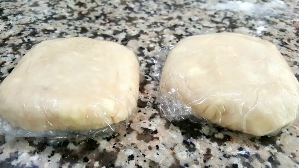 two unbaked flat balls of pie dough each wrapped in plastic wrap
