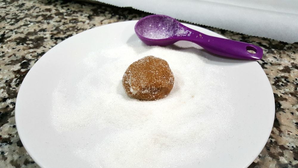 ginger snap cookie dough ball rolled in sugar sitting on a plate of sugar with a purple tablespoon