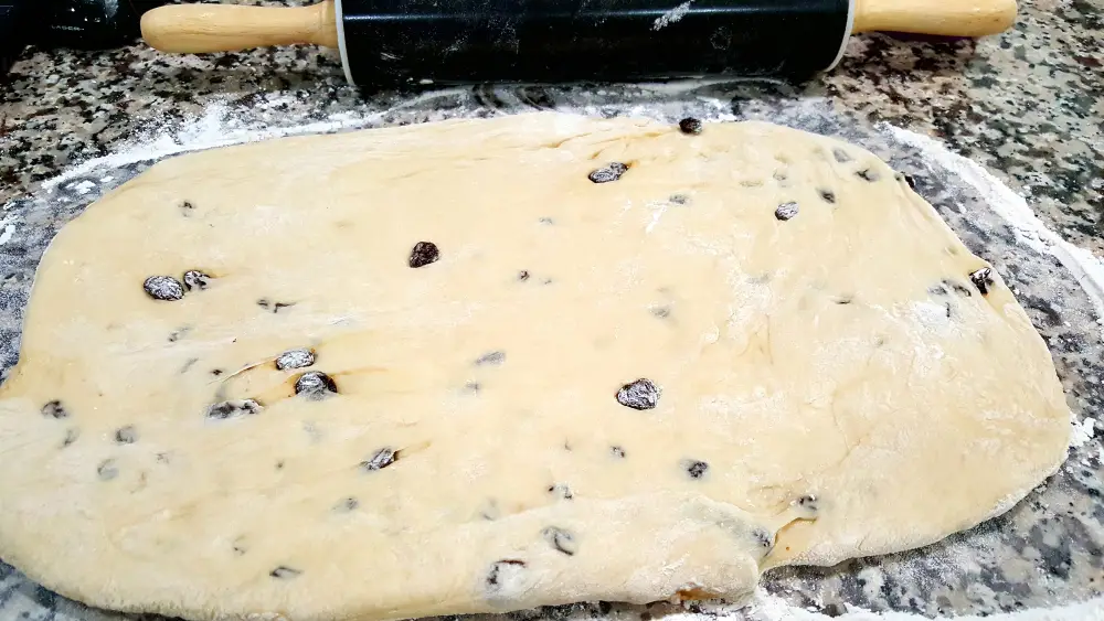 raisin bread dough with raisins in it rolled out into a rectangle with a rolling pin.