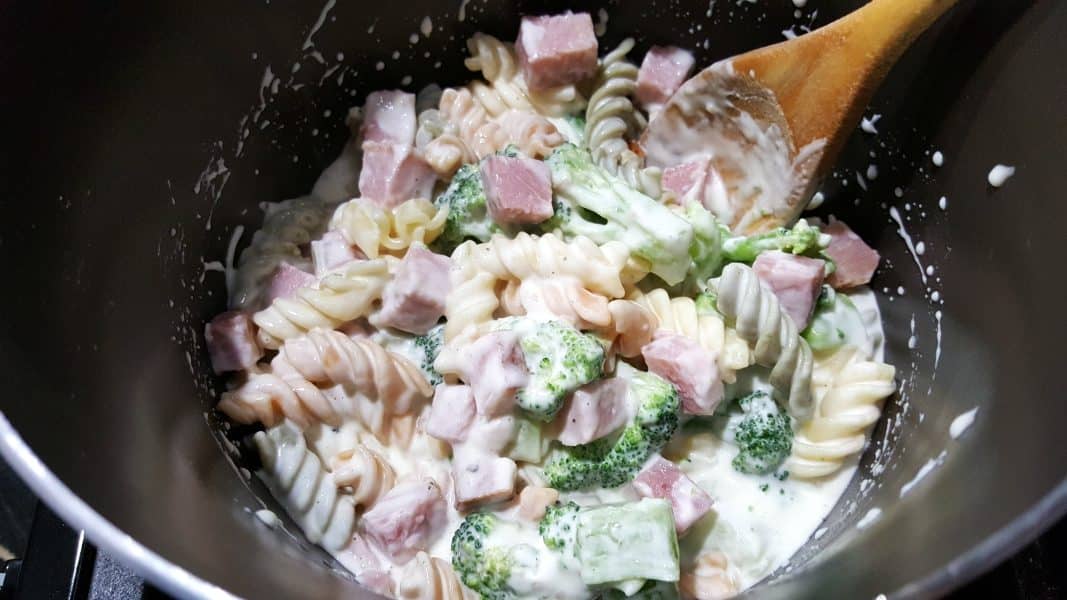 cheese sauce, pasta, broccoli, and ham in a sauce pan with a wooden spoon