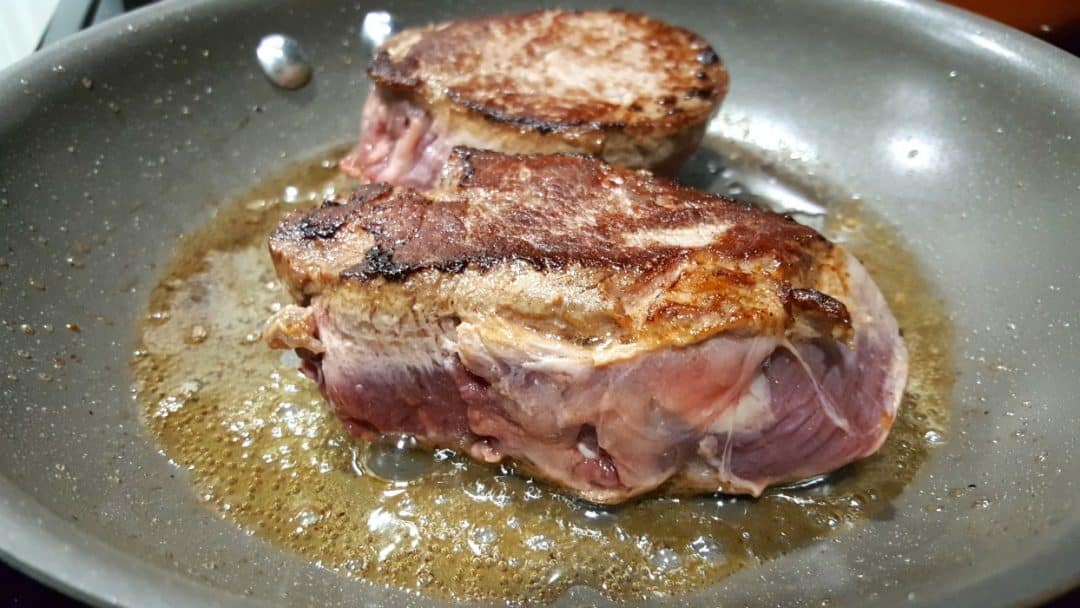 two beef tenderloin steaks cooking in a pan.