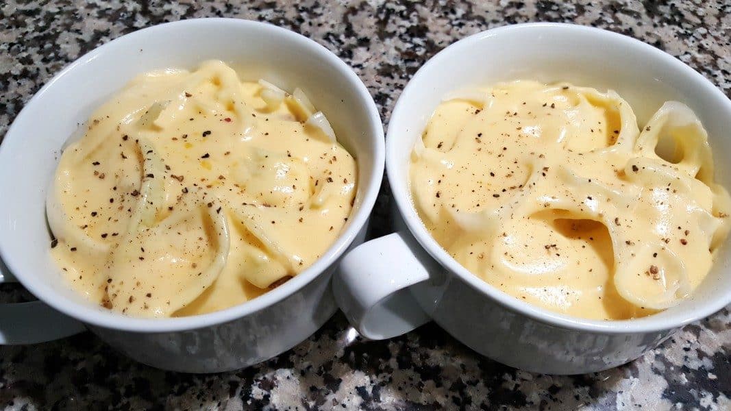 two bowls filled with sliced potatoes, sliced onions, and topped with cheese sauce and black pepper