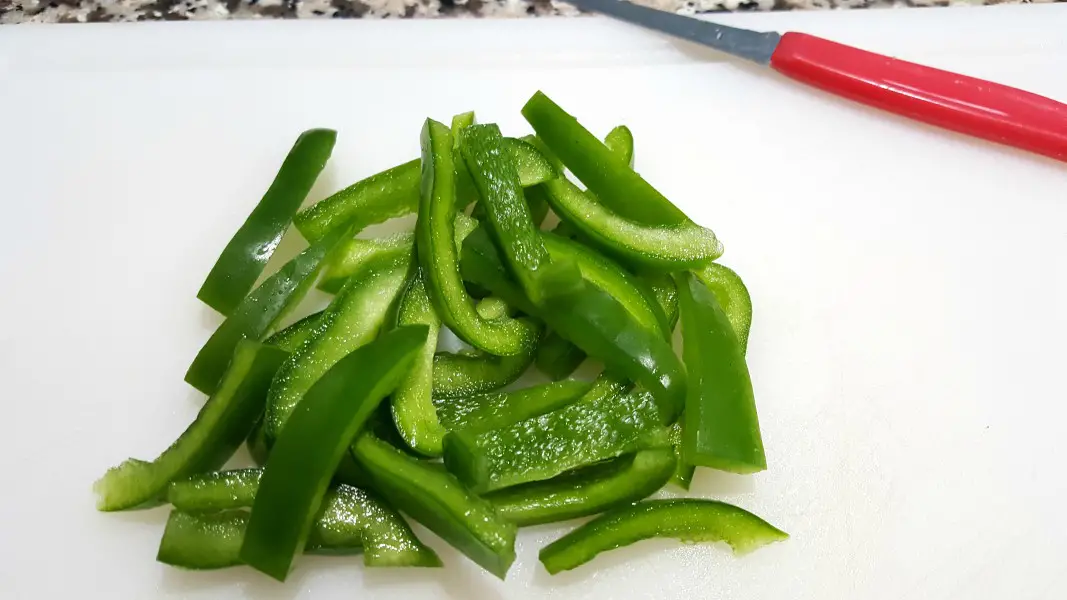 green peppers sliced thin on a cutting board with a red pairing knife.