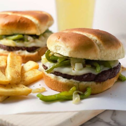 a close up of two philly cheesesteak burgers and pile of seasoned fries on white paper on a cutting board with a glass of beer in the back