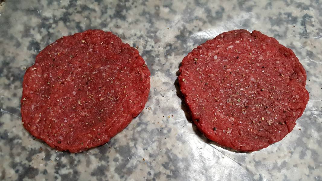 two perfectly round shaped raw beef hamburgers sprinkled with salt and pepper.
