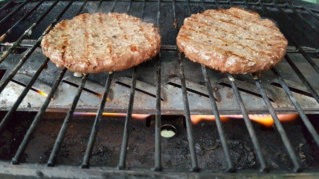 two round ground beef quarter pound patties cooking over a flame on a grill
