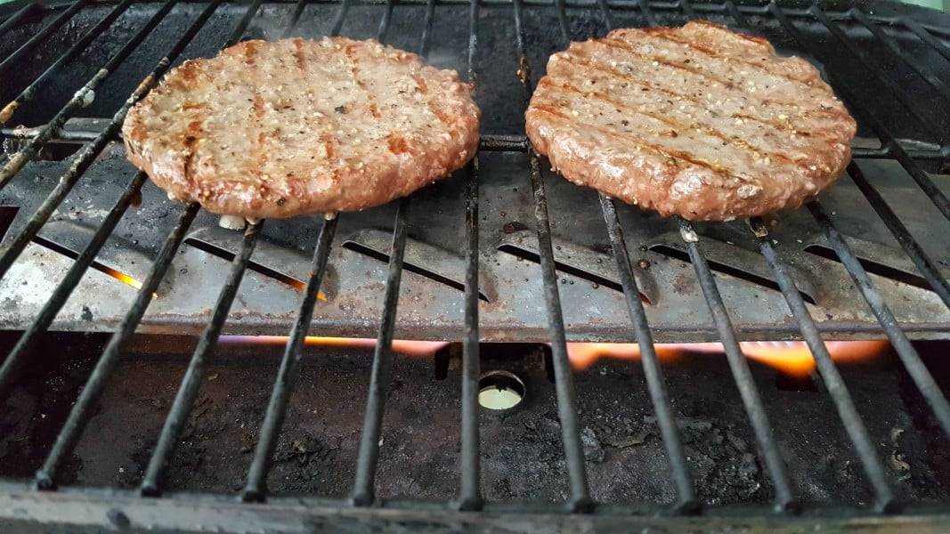 two round ground beef quarter pound hamburger patties cooking over a flame on a grill.