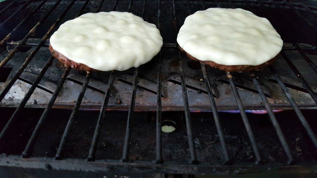 two round ground beef quarter pound patties cooking over a flame on a grill with melted white cheese on top