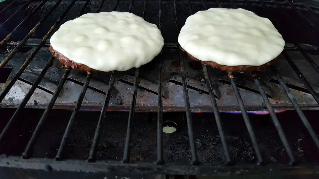two round ground beef quarter pound patties cooking over a flame on a grill with melted white cheese on top.