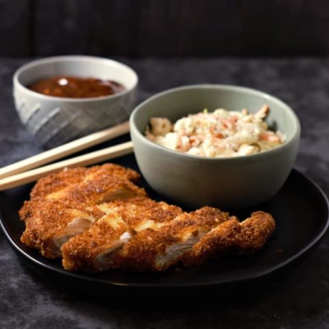 a plate with a breaded pork loin, chopsticks, and a bowl of coleslaw on it, and a bowl on the side with brown sauce in it