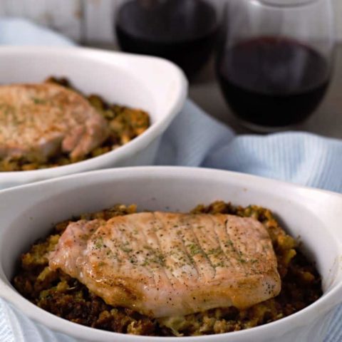 a close up of two baking dishes filled with stuffing topped with a pork chop next to two glasses of wine