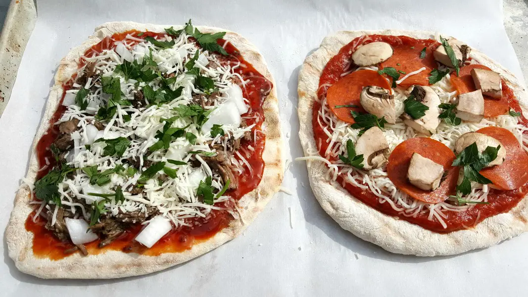 two uncooked pizzas with toppings added ready to grill.