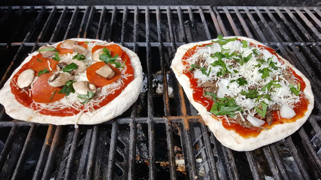 two pizzas with toppings cooking on a grill.