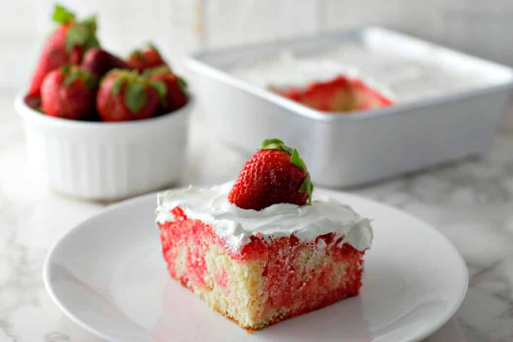 a slice of Easy Homemade Strawberry Poke Cake with Jello with rest of cake still in the pan and a bowl of strawberries.