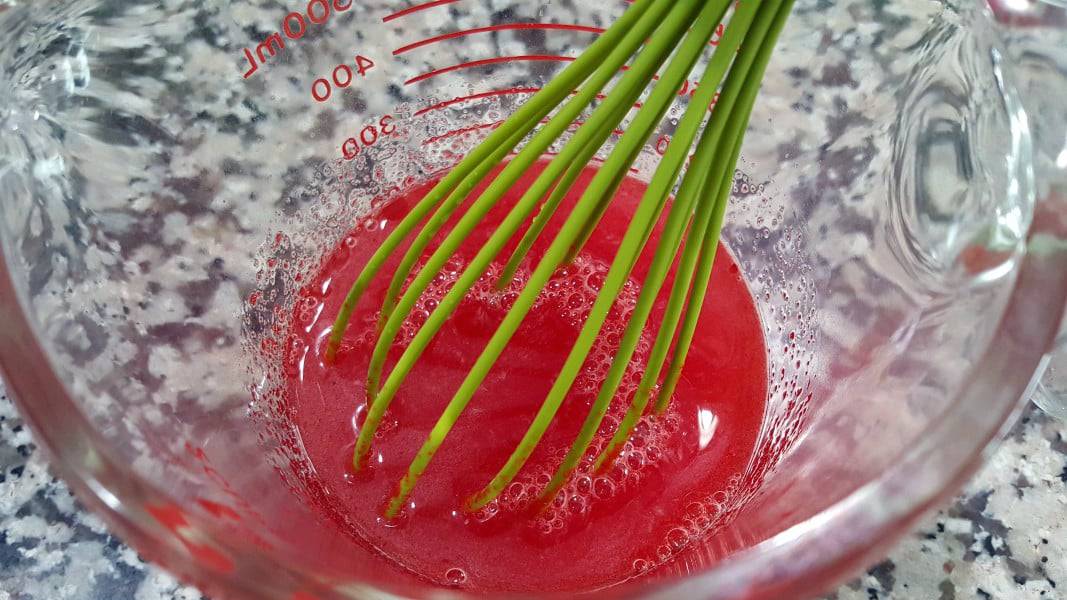 strawberry jello and hot water mixed in a measuring cup with a whisk.