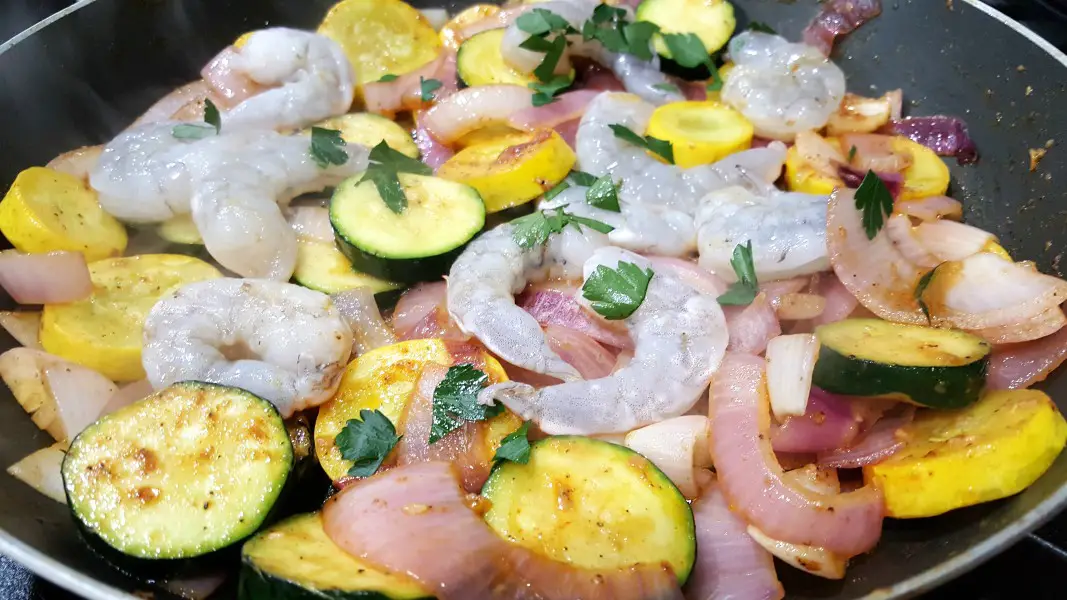 zucchini, yellow squash, and red onion mixed with garlic, olive oil, paprika, salt and pepper cooking in a skillet with shrimp and parsley.