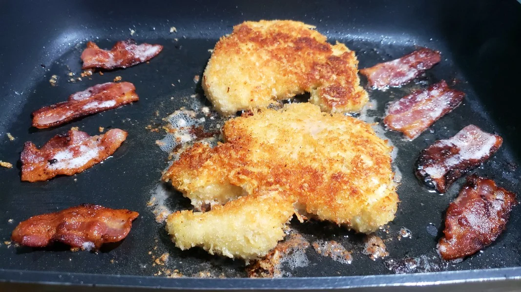 two pieces of parmesan breaded chicken frying in a pan with eight half slices of bacon.
