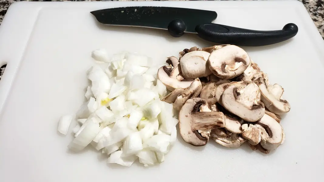 sliced mushrooms and diced onions on a cutting board with a knife.