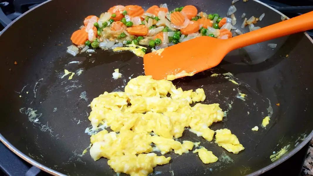 scrambled eggs cooking in a pan with carrots, peas, onions, and garlic.