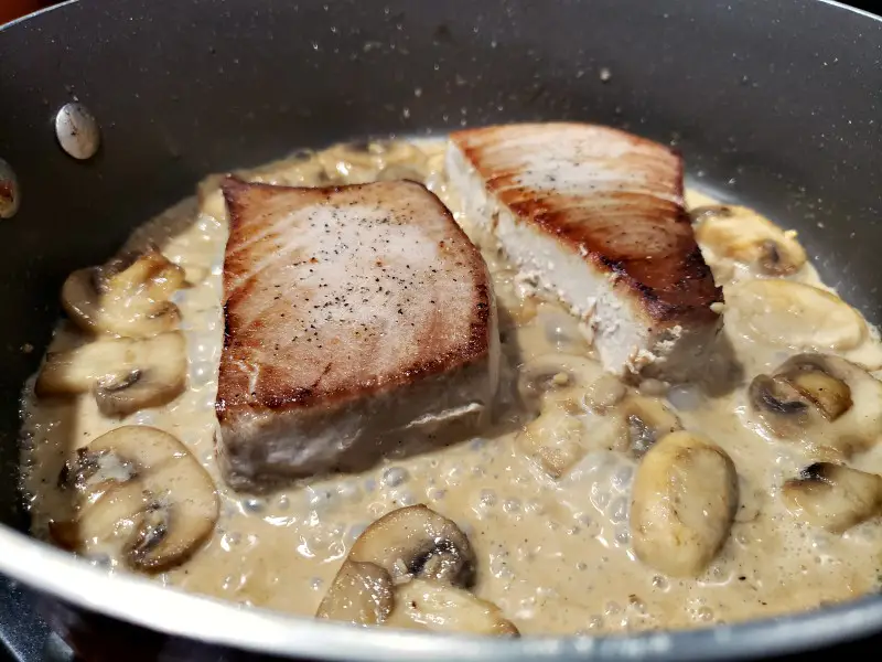 two tuna steaks cooking in a pan with creamy mushroom sauce
