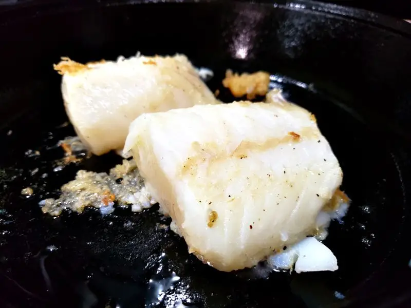 2 cod fillets cooking in a cast iron skillet