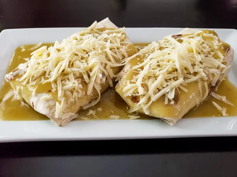 two cheesy smothered pulled pork burritos covered in green sauce and white shredded cheese on a platter.