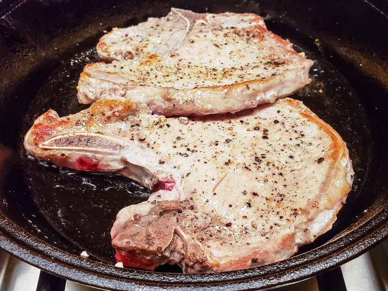 two pork chops cooking in a cast iron skillet.