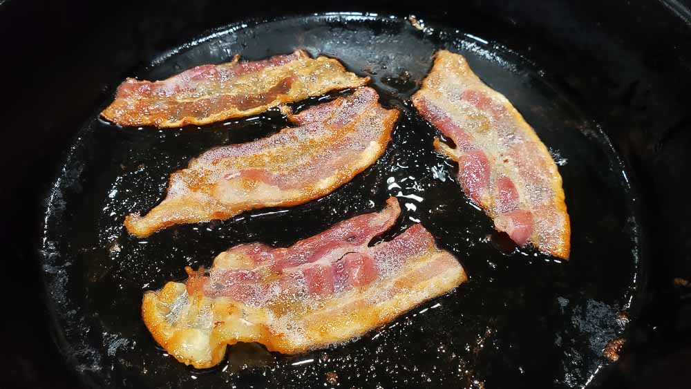 4 halves of bacon frying in a cast iron skillet.