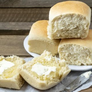 three dinner rolls stacked on a plate and one dinner roll cut open is spread with butter