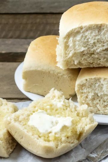 three dinner rolls stacked on a plate and one dinner roll cut open is spread with butter