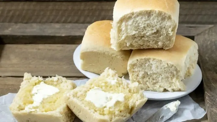 three dinner rolls stacked on a plate and one dinner roll cut open is spread with butter