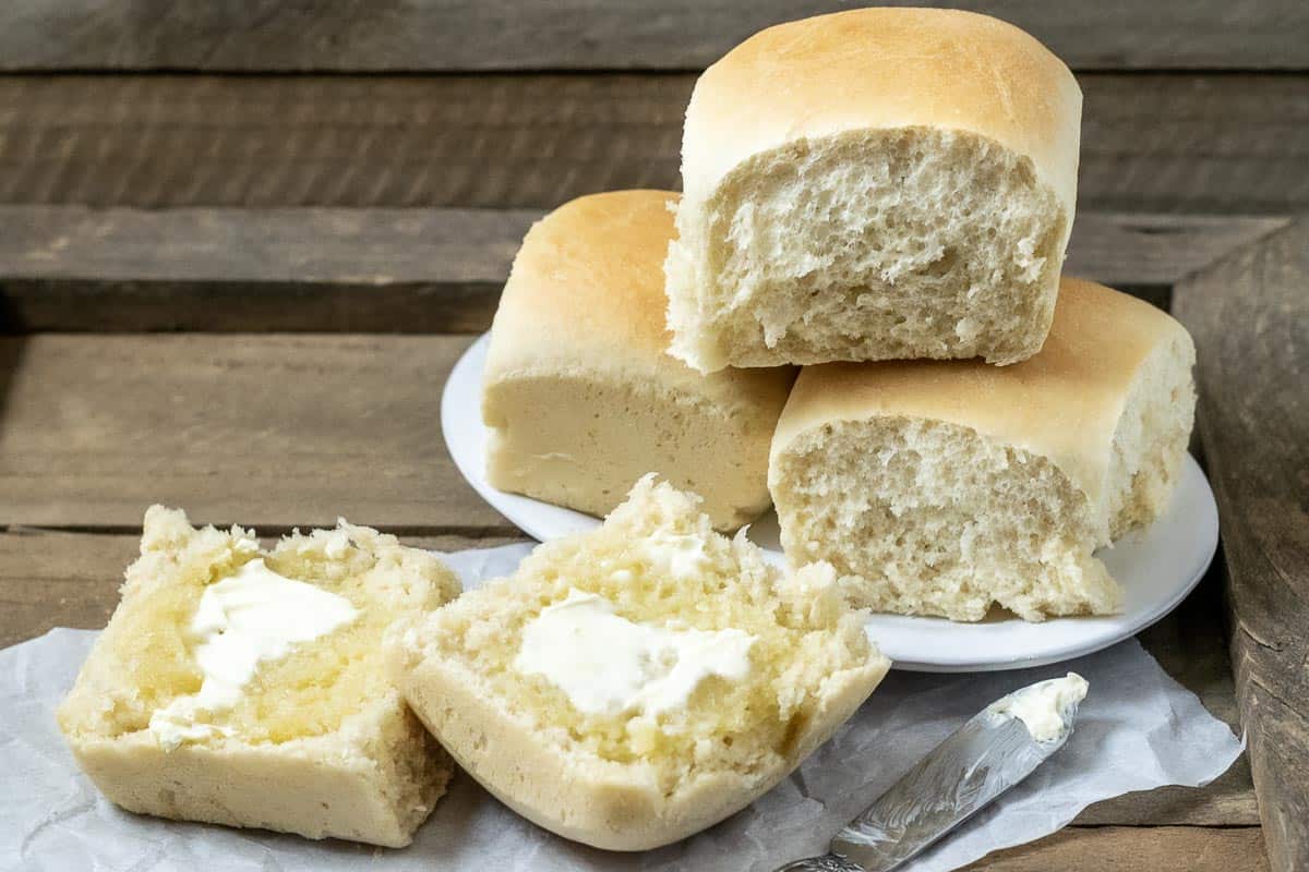 three Old Fashioned Dinner Rolls stacked on a plate and one dinner roll cut open is spread with butter.