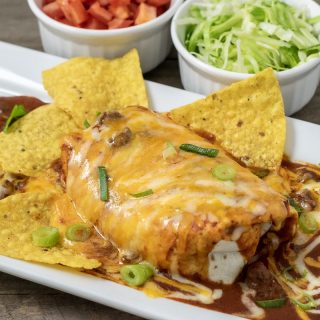 a large wet burrito on a platter with tortilla chips and sides of diced tomato and shredded lettuce