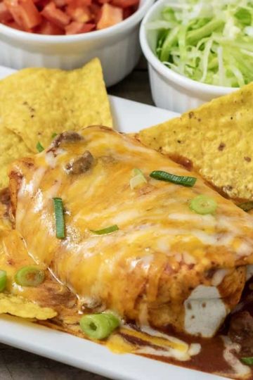 a large wet burrito on a platter with tortilla chips and sides of diced tomato and shredded lettuce