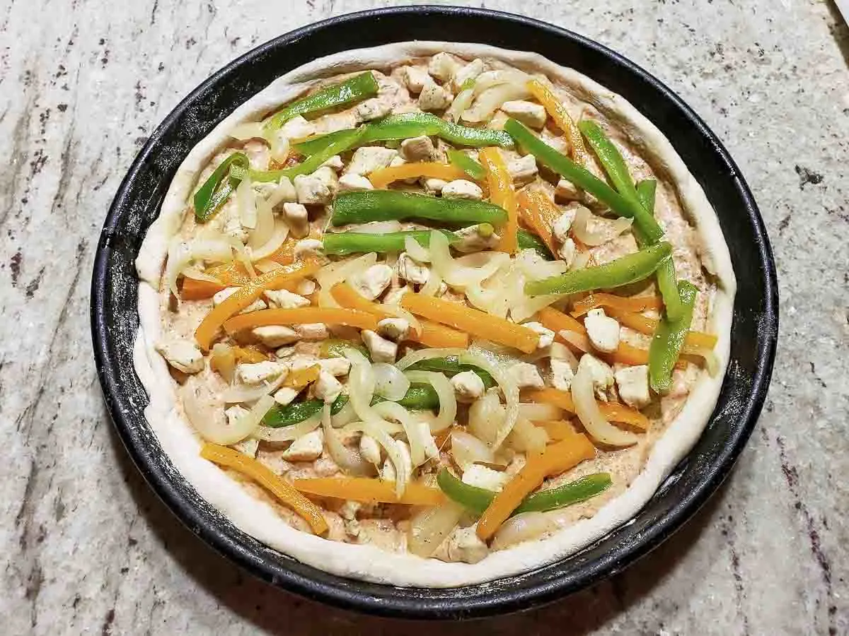 sliced fajita veggies and chicken layered on top of sauce in pizza pan.