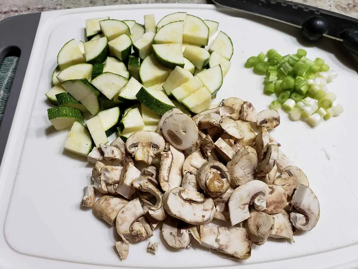 chopped zucchini, mushrooms, and green onion on a white cutting board.