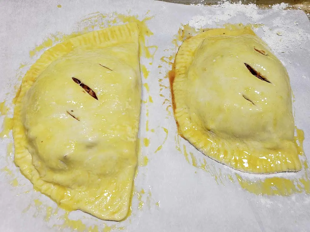 two filled pastries on a baking tray brushed with beaten egg and slits cut in the top.
