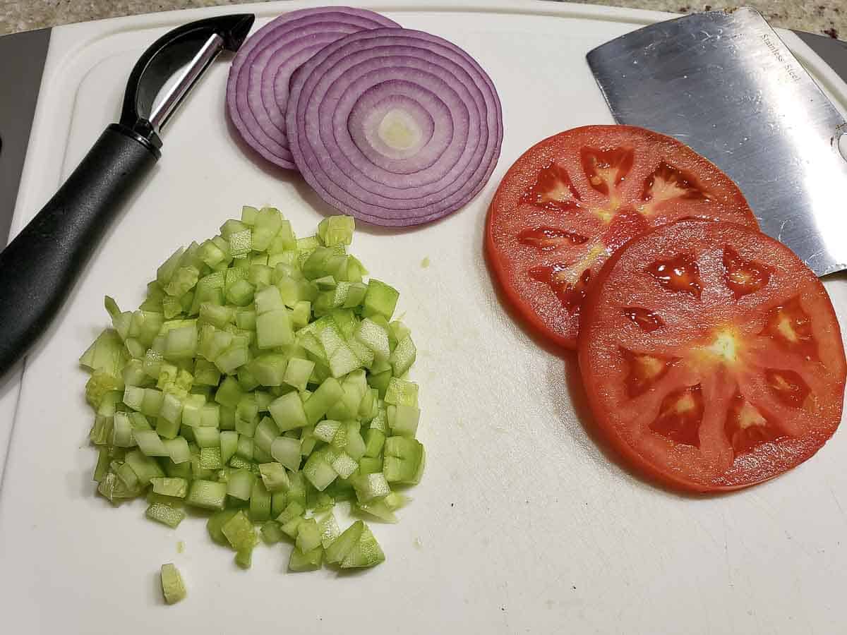 diced cucumber, sliced red onion, and sliced tomato on a cutting board.