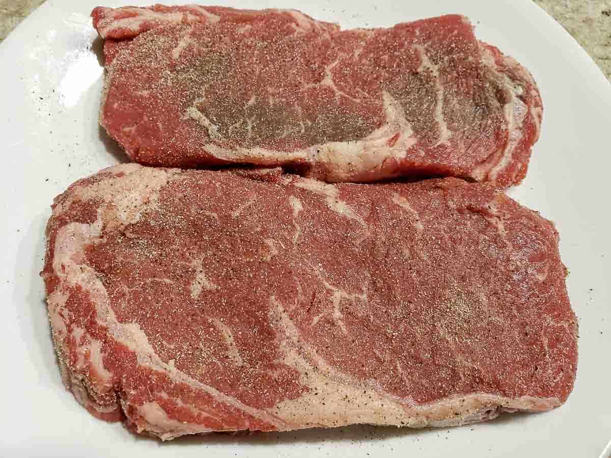 two ribeye steaks seasoned with salt and pepper on a plate.