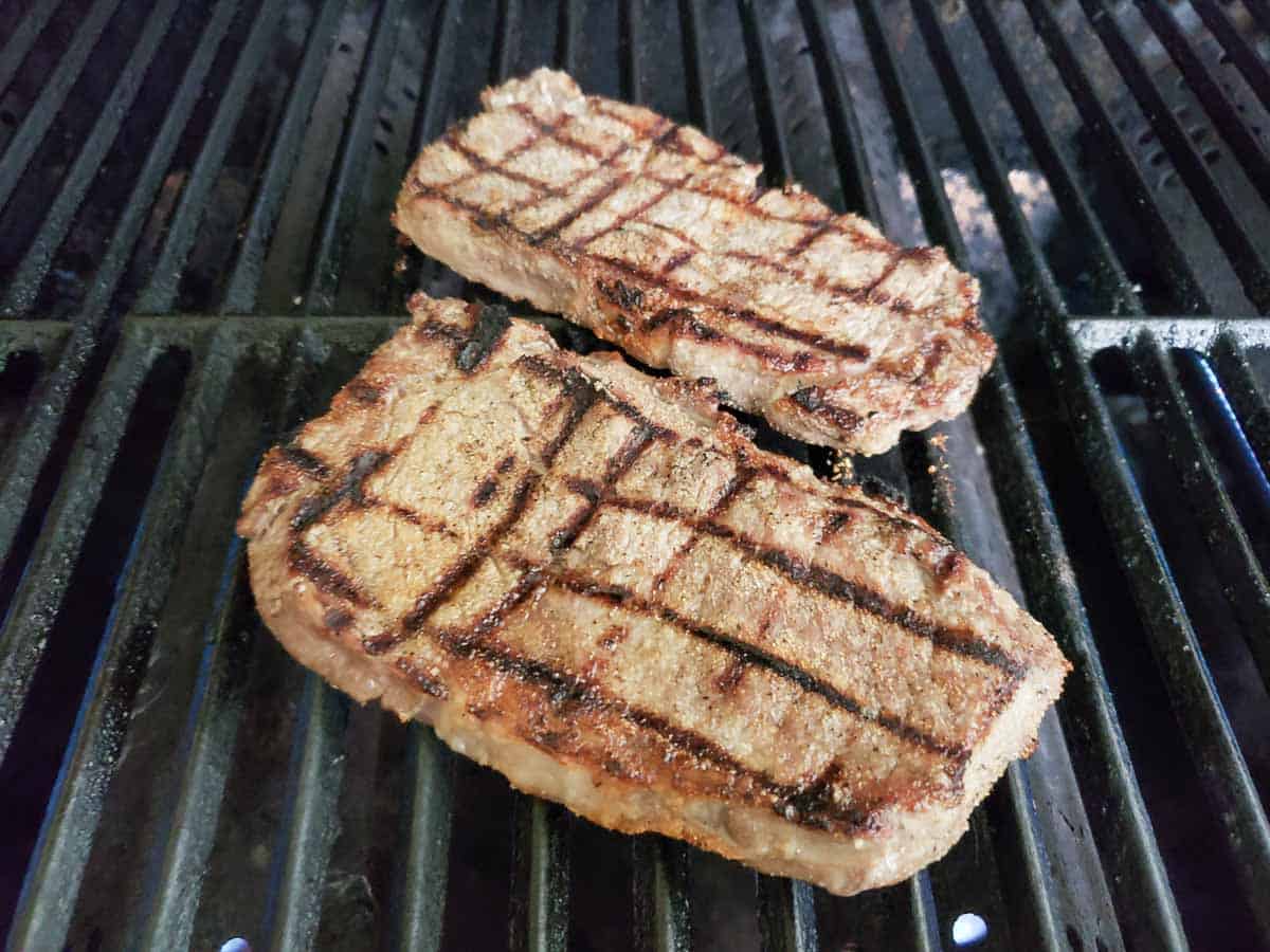 two ribeye steaks cooking on a gas grill.