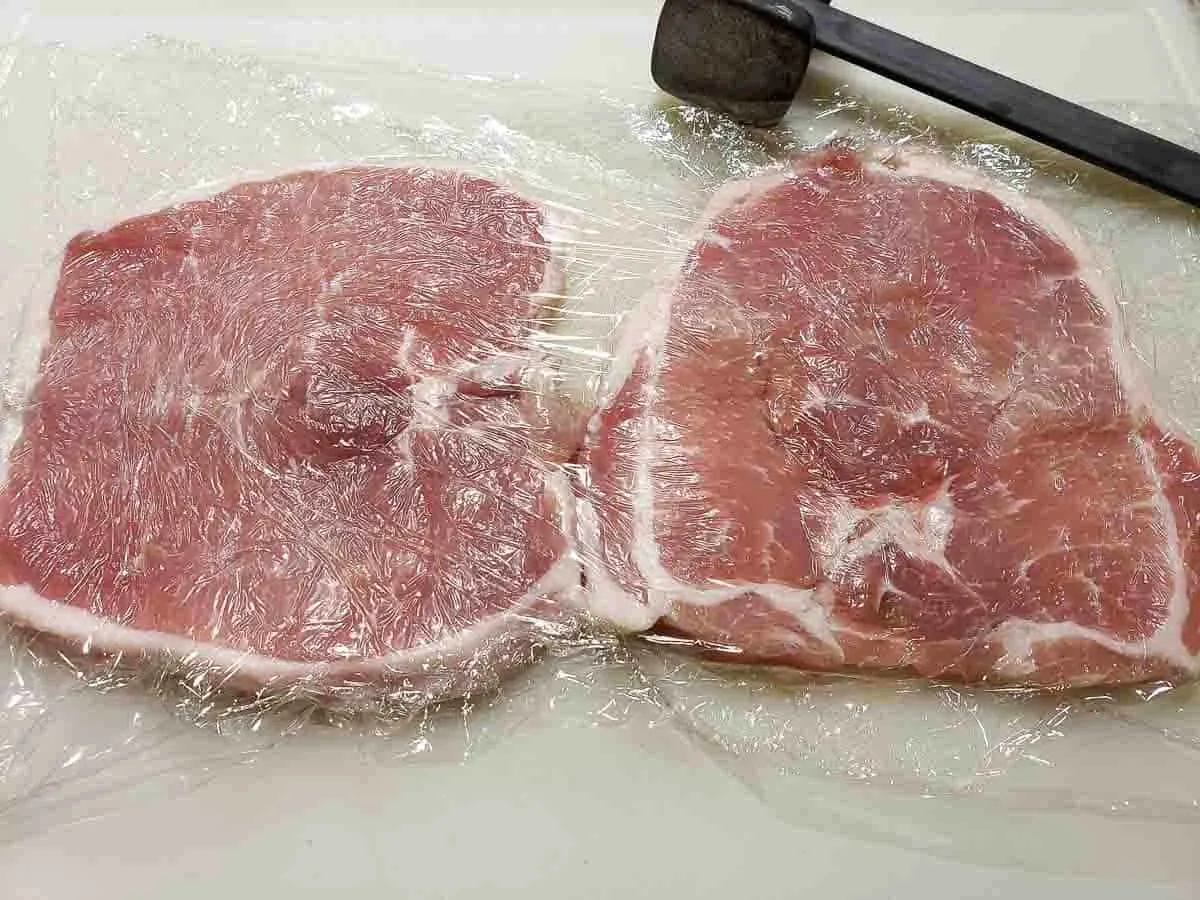 two bonless pork chops pounded flat between sheets of plastic wrap.
