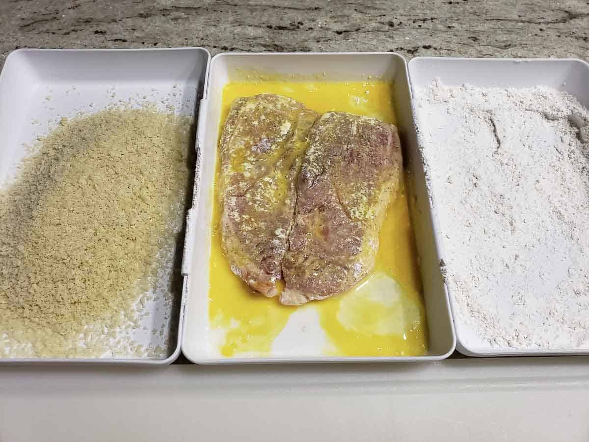 3 dishes side by side with Panko bread crumbs, a beaten egg with a pork chops in it, and flour.