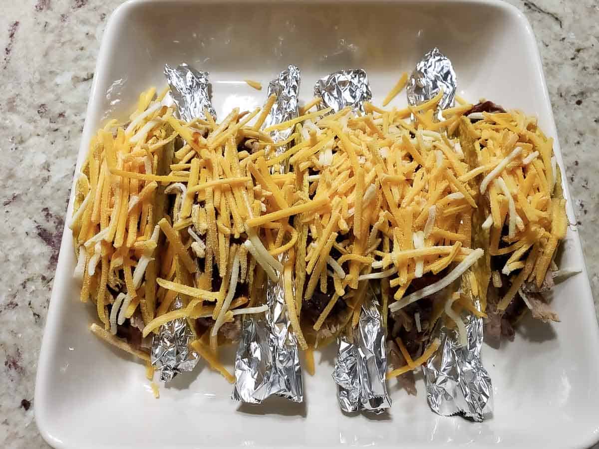 5 taco shells filled with pulled pork, BBQ sauce, and cheese and rolled tinfoil in between in a baking dish