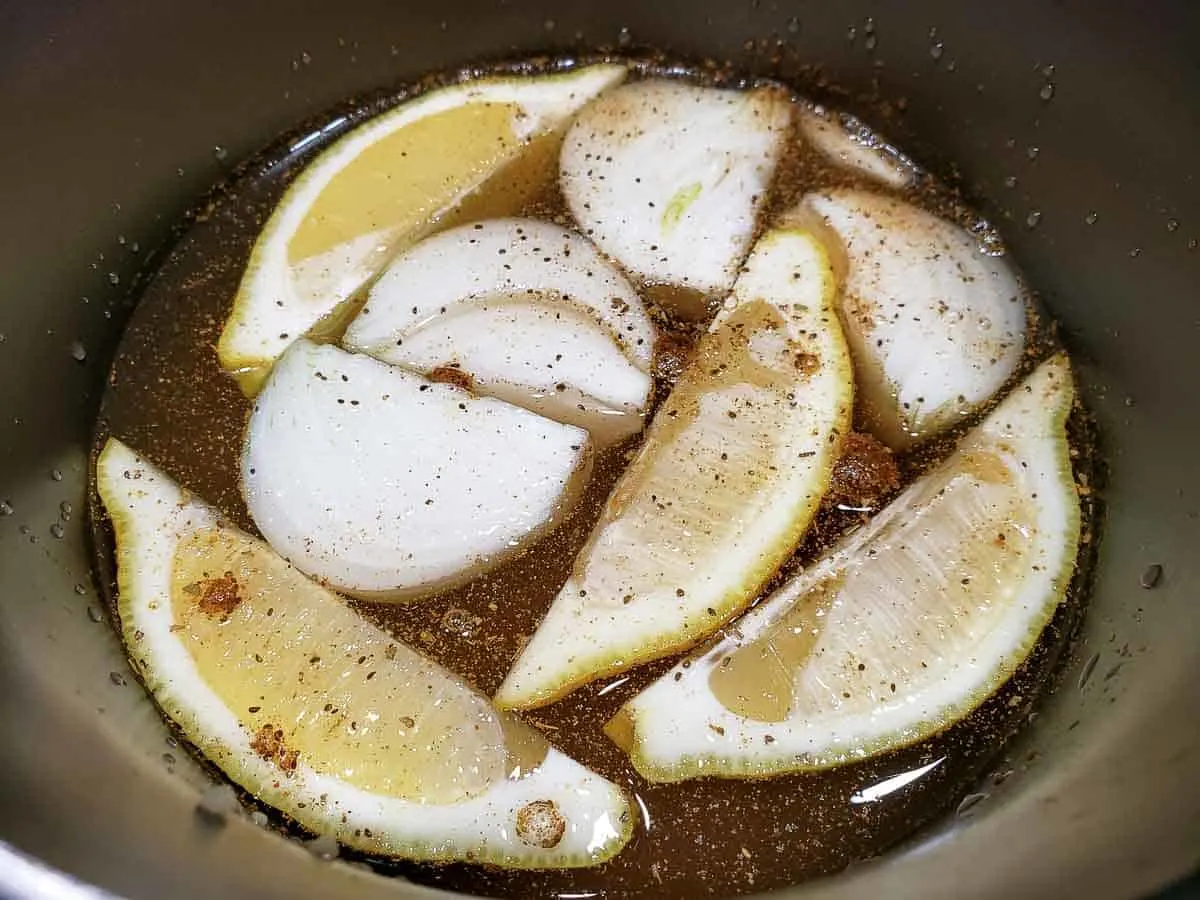 a pan filled with brown liquid, seasonings, onions, and lemon wedges.