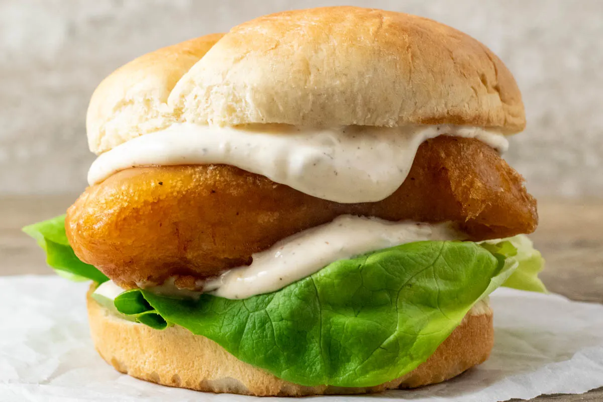 up close front view of a bun filled with lettuce, white creamy sauce, and golden brown fish filet