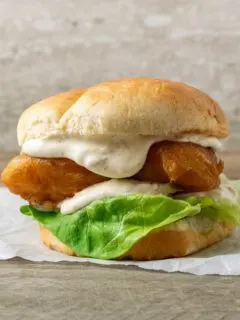 a bun filled with lettuce, white creamy sauce, and golden brown fish filet on parchment paper.