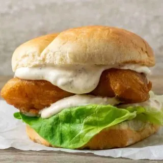 a bun filled with lettuce, white creamy sauce, and golden brown fish filet on parchment paper.